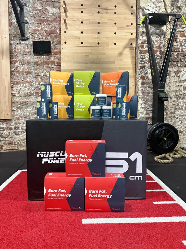 Gestapelte Sport Nutrition Boxen mit der Aufschrift „Repeat The Switch“ und „Spark your energy!“ in einem Fitnessstudio mit Hanteln und Trainingsgeräten im Hintergrund.